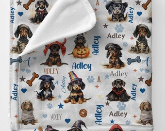 May include: A white fleece blanket featuring a repeating pattern of dachshund puppies in various costumes, including a skeleton, a pumpkin, a wizard, and a pirate. The name Adley is printed in blue and brown text alongside paw prints and bones.