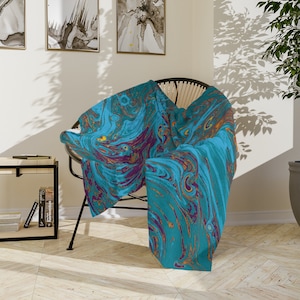 May include: A teal blue throw blanket with a swirling abstract pattern in shades of purple, gold, and brown. The blanket is draped over a wicker chair in a modern living room setting.