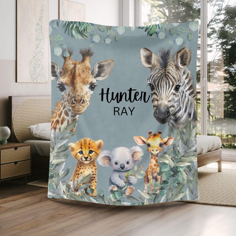 May include: A soft blanket featuring a light blue background with illustrations of a giraffe, a zebra, a cheetah cub, a koala, and a small giraffe. The text "Hunter RAY" is printed in black in the center, surrounded by green eucalyptus leaves.