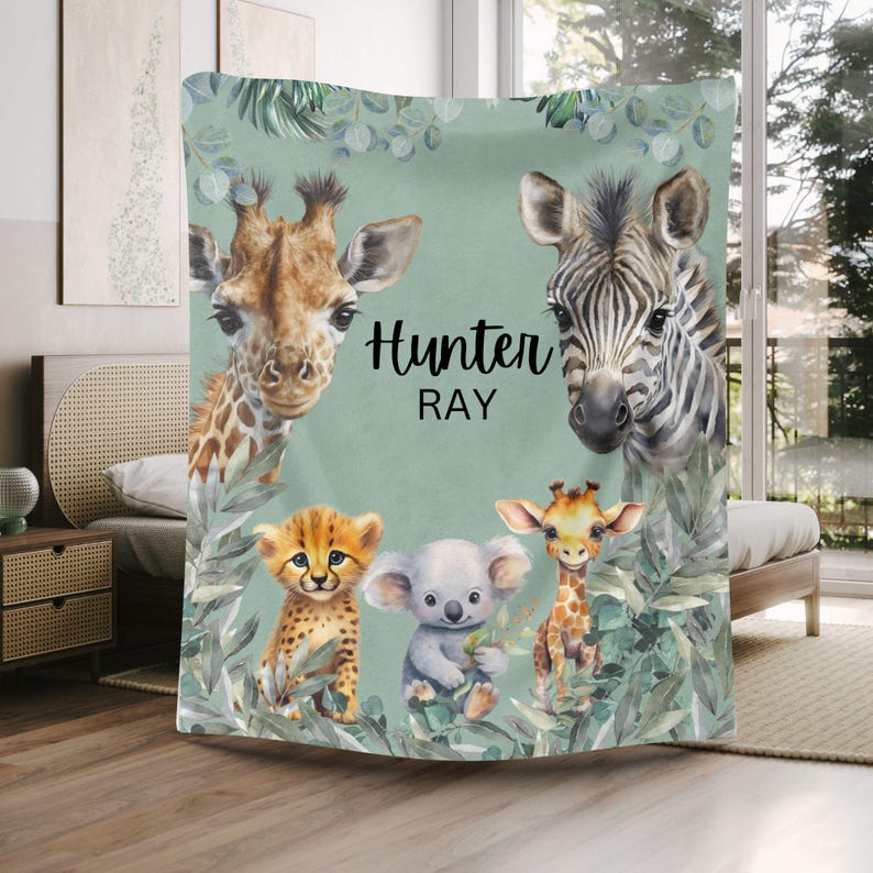 May include: A soft blanket featuring a light green background with illustrations of a giraffe, a zebra, a cheetah cub, a koala, and a small giraffe. The text Hunter RAY is printed in black in the center, surrounded by green foliage and leaves.