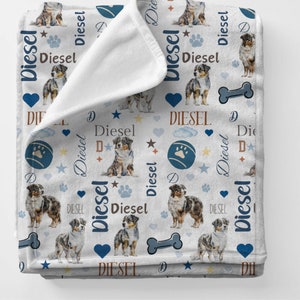Puede incluir: Manta de franela blanca con un patrón repetido de un perro pastor australiano azul y marrón, una pelota azul con una huella de pata, un hueso, un corazón, una estrella y la palabra "Diesel" en varias fuentes y colores.