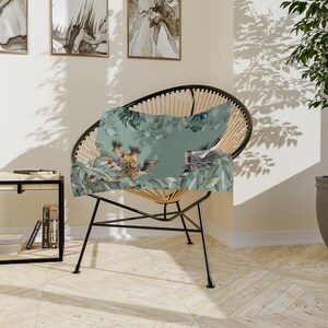 May include: A rectangular throw pillow featuring a printed illustration of a giraffe and a zebra head nestled among green tropical leaves and eucalyptus branches on a soft sage green background, resting on a black wire frame accent chair.
