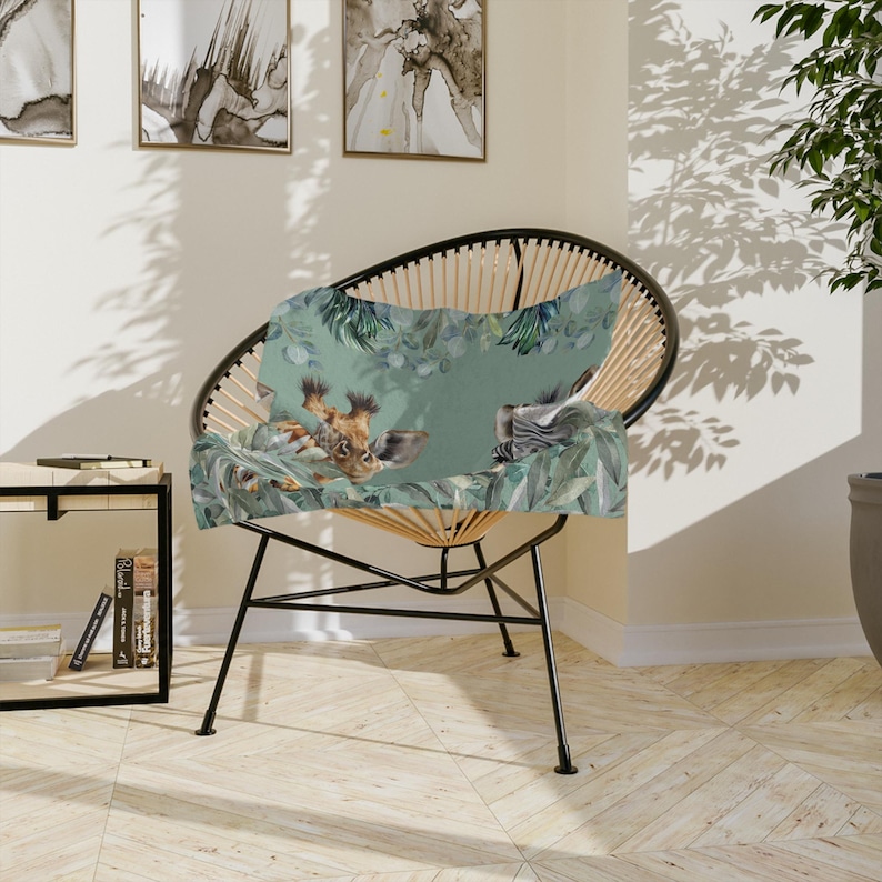 May include: A rectangular throw pillow featuring a printed illustration of a giraffe and a zebra head nestled among green tropical leaves and eucalyptus branches on a soft sage green background, resting on a black wire frame accent chair.