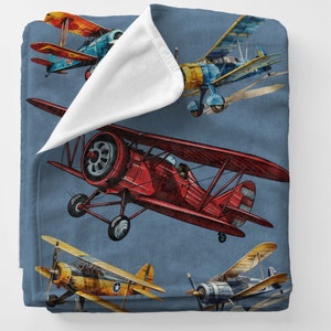 May include: A blue blanket featuring watercolor illustrations of vintage biplanes in red, yellow, and blue. The biplanes have propellers and are set against a blue background. The blanket has a soft, white underside.