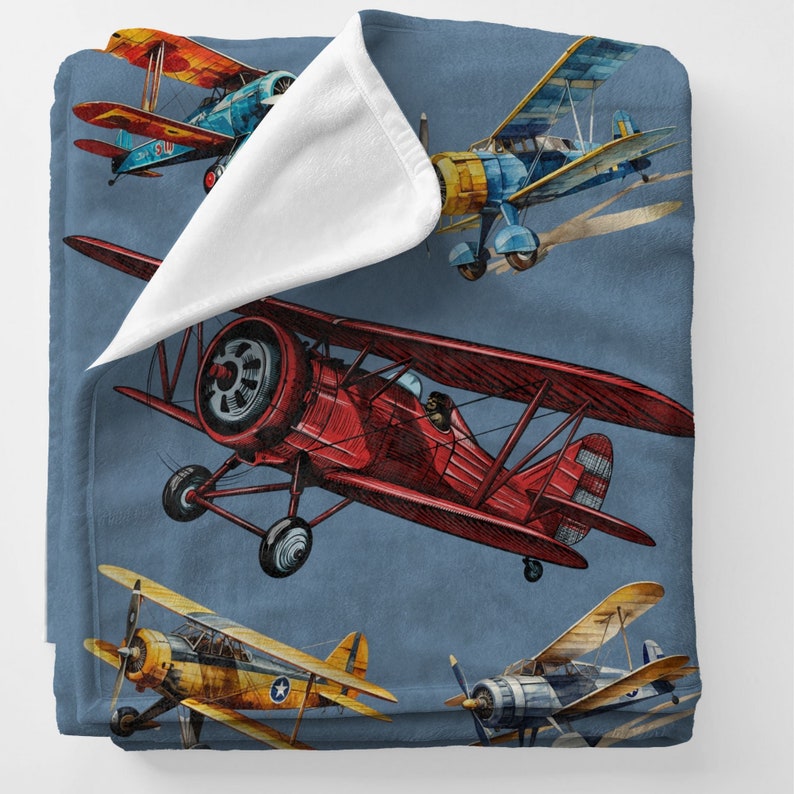 May include: A blue blanket featuring watercolor illustrations of vintage biplanes in red, yellow, and blue. The biplanes have propellers and are set against a blue background. The blanket has a soft, white underside.