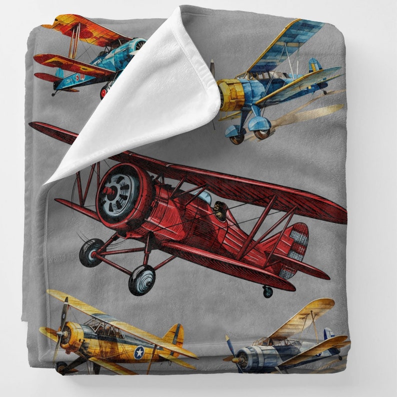 May include: A gray blanket featuring a watercolor illustration of vintage biplanes in shades of red, blue, yellow, and orange. The biplanes are depicted in flight against a gray background. The blanket has a soft, plush texture.