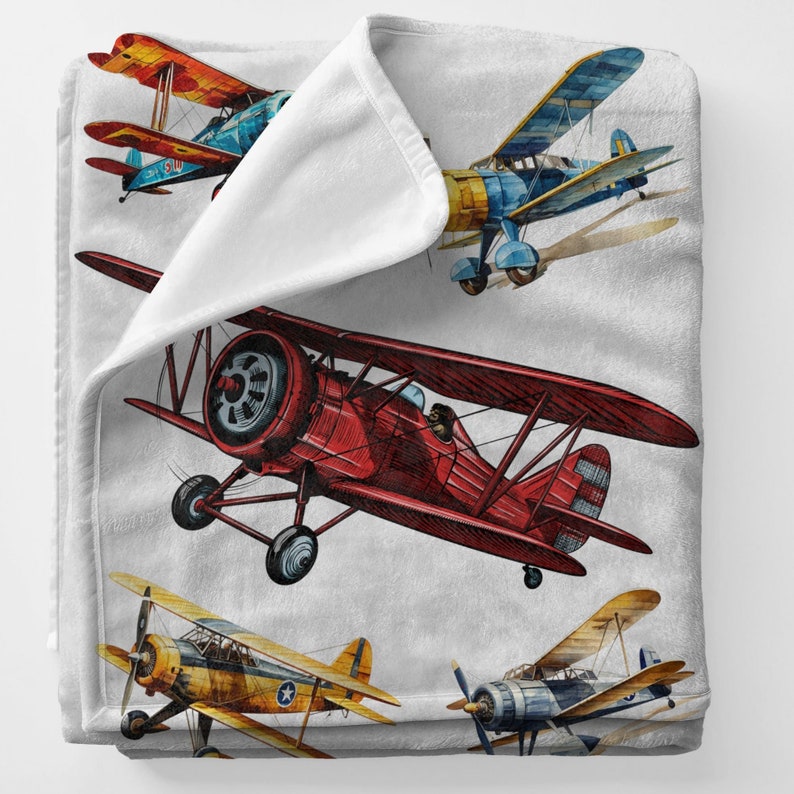 May include: A soft, white blanket featuring a watercolor design of vintage biplanes in shades of red, blue, yellow, and orange. The blanket has a folded edge, showcasing the soft, plush material.