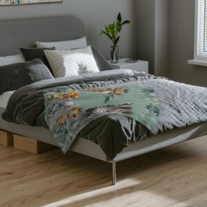 May include: A soft throw blanket featuring a repeating pattern of illustrated safari animals including a zebra, giraffe, and cheetah set against a muted sage green background with green foliage accents, draped over a gray bed frame.