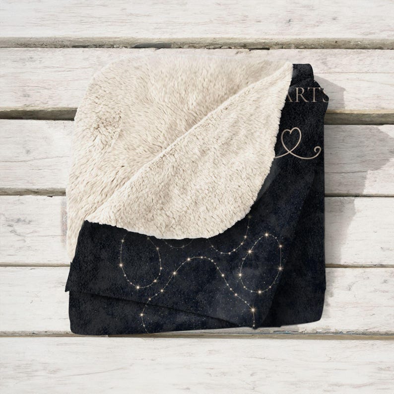 May include: A black and cream-colored blanket with a constellation design. The blanket has a soft, plush cream-colored interior and a dark exterior with a pattern of connected dots. The word "ARTS" and a heart are visible on the blanket.
