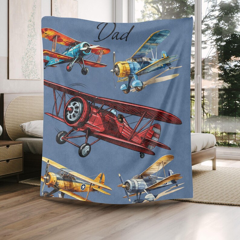 May include: A blue blanket featuring watercolor illustrations of vintage airplanes in shades of red, yellow, blue, and orange. The word "Dad" is written in cursive at the top. This blanket is perfect for aviation enthusiasts.