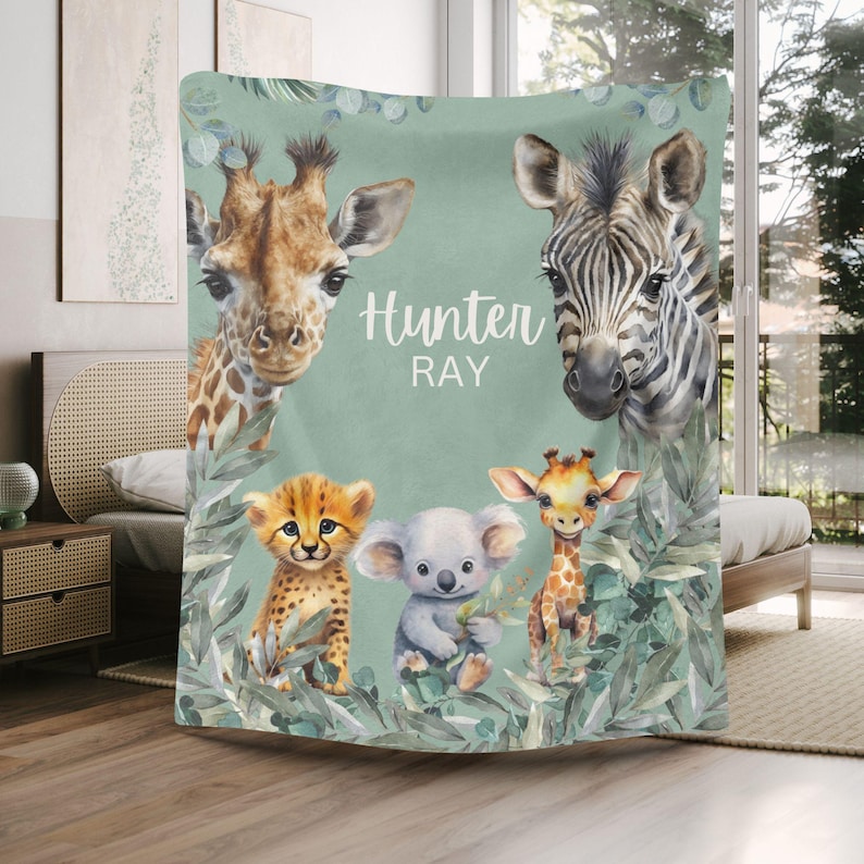 May include: A soft blanket featuring a sage green background with watercolor illustrations of a giraffe, a zebra, a cheetah cub, a koala, and a smaller giraffe. The text "Hunter RAY" is printed in white in the center, surrounded by eucalyptus leaves.
