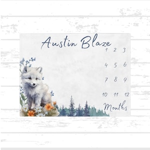 Op de afbeelding: Een witte babygroeikaart met een aquarelillustratie van een witte poolvos in een bosrijke omgeving. De kaart is gelabeld "Austin Blaze" en heeft de nummers 1 tot en met 12 voor elke maand van het jaar. De kaart is op een witte houten achtergrond.