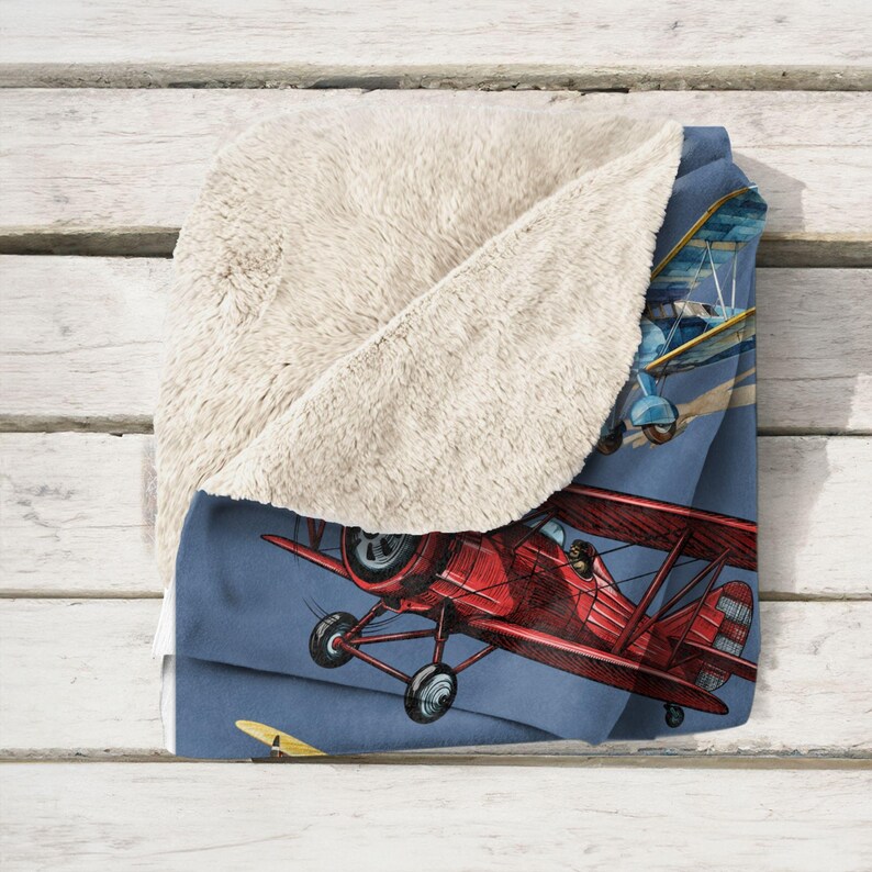 May include: A soft, plush blanket featuring a vintage airplane design. The blanket has a blue background with a red biplane in flight, and a cream-colored sherpa lining for added warmth and comfort.