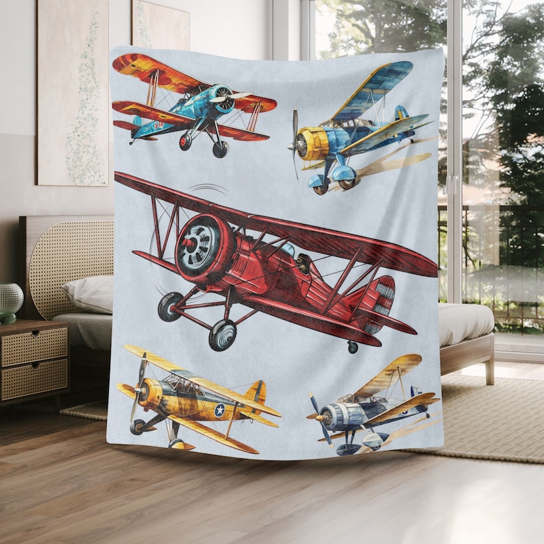 May include: A soft, light blue blanket featuring a watercolor illustration of five vintage biplanes in flight. The planes are in various colors, including red, yellow, blue, and orange, with detailed propeller and wing designs.