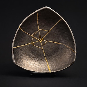 May include: A dark gray, triangular ceramic bowl with gold-colored kintsugi repairs. The bowl has a metallic sheen and is displayed against a black background. The cracks are filled with gold, highlighting the history of the piece.