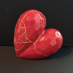 May include: A glossy red heart-shaped sculpture with gold-colored lines. The heart has a faceted surface and a smooth, reflective finish. The gold lines create a visual representation of the Japanese art of Kintsugi.