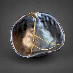 May include: A small, irregularly shaped ceramic bowl with a dark, metallic, and textured finish. The bowl features a prominent repair line running through its center, highlighted with a bright gold-colored lacquer in the kintsugi style.