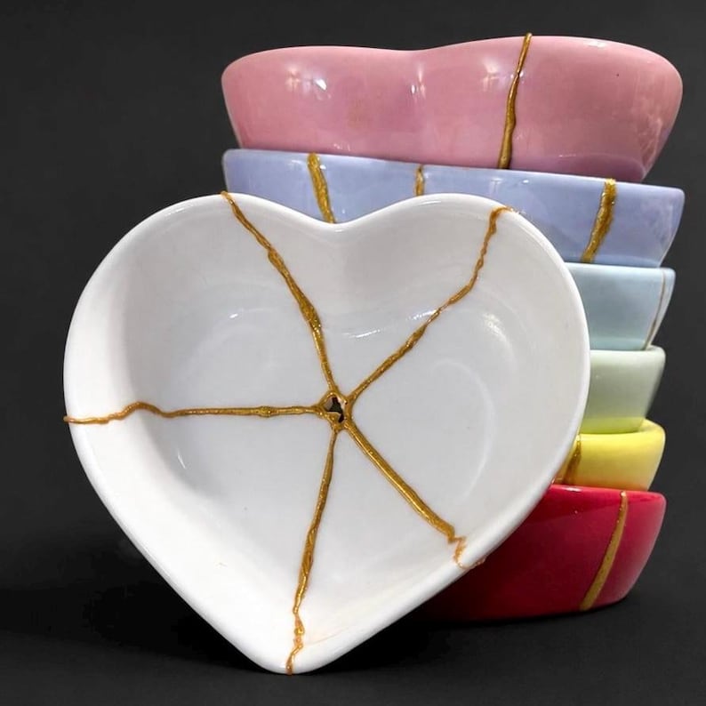 May include: A stack of six heart-shaped ceramic bowls in various pastel colours including pink, light blue, light green, yellow, and red. Each bowl features a white interior with a gold-coloured crack pattern radiating from the centre.