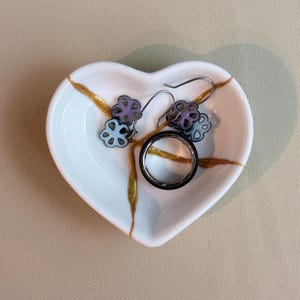 May include: A white heart-shaped ceramic dish featuring gold-coloured cracks across the surface. Inside the dish are a pair of dangling earrings with light purple and blue flower-shaped charms and a smooth, dark metallic ring with a silver band.