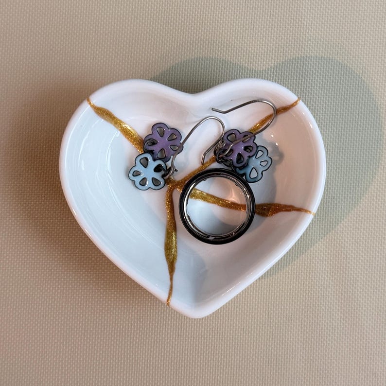 May include: A white heart-shaped ceramic dish featuring gold-coloured cracks across the surface. Inside the dish are a pair of dangling earrings with light purple and blue flower-shaped charms and a smooth, dark metallic ring with a silver band.