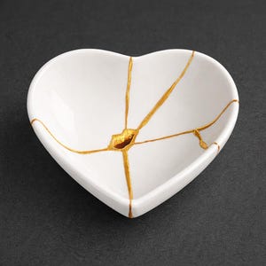 May include: A white heart-shaped ceramic dish with gold-colored kintsugi-style repairs. The dish has a smooth, glossy finish and is set against a dark background. The gold lines highlight the cracks, creating a unique decorative piece.