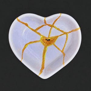 May include: A heart-shaped ceramic dish in a pale lavender hue, featuring a network of cracks repaired with a metallic gold finish. The gold lines radiate from a central point, creating a web-like pattern across the smooth, glossy surface.