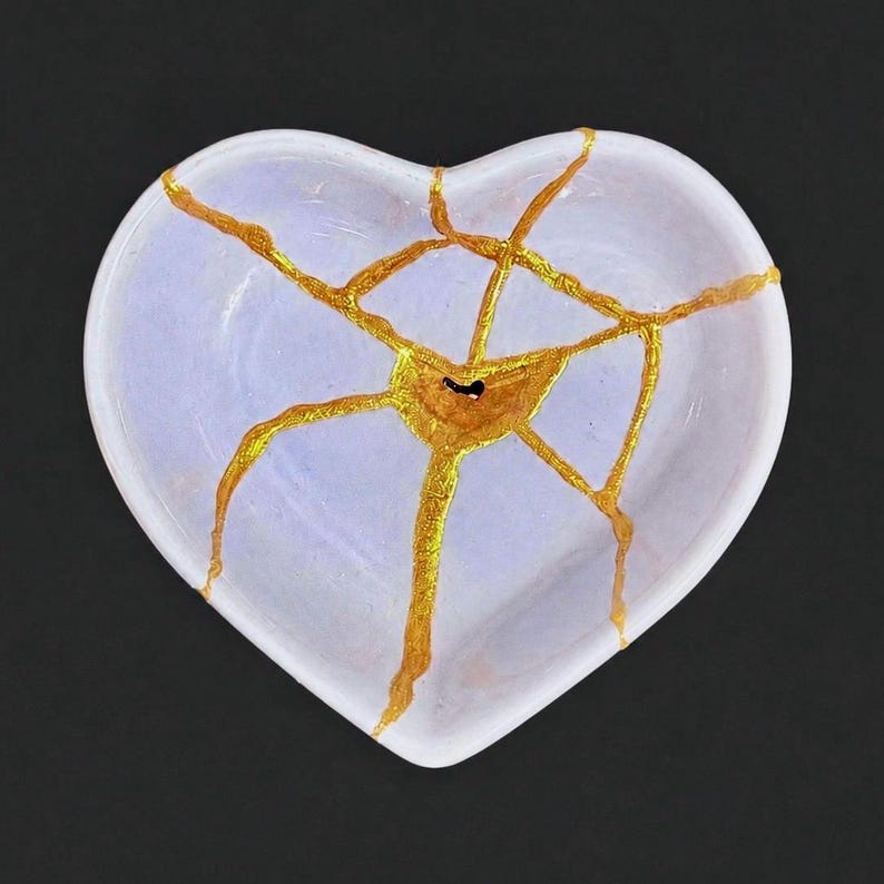 May include: A heart-shaped ceramic dish in a pale lavender hue, featuring a network of cracks repaired with a metallic gold finish. The gold lines radiate from a central point, creating a web-like pattern across the smooth, glossy surface.