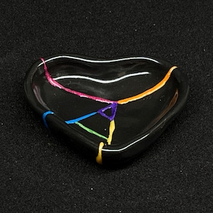 May include: A heart-shaped black ceramic dish with colorful, visible cracks. The cracks are filled with pink, blue, green, orange, and yellow paint. The dish has a glossy finish and is set against a dark background.