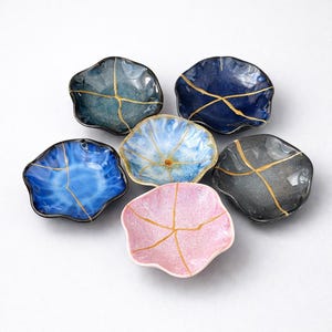 May include: A set of six small, scalloped-edge ceramic dishes in various colors including blue, pink, gray, and teal. Each dish features a distinct gold-colored line pattern across the surface, resembling the kintsugi repair technique.