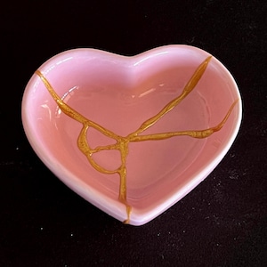 May include: A heart-shaped ceramic dish in a soft pastel pink colour. The interior surface features a prominent gold-coloured vein pattern that resembles a repair line, extending across the centre of the bowl from the top edge to the bottom point.