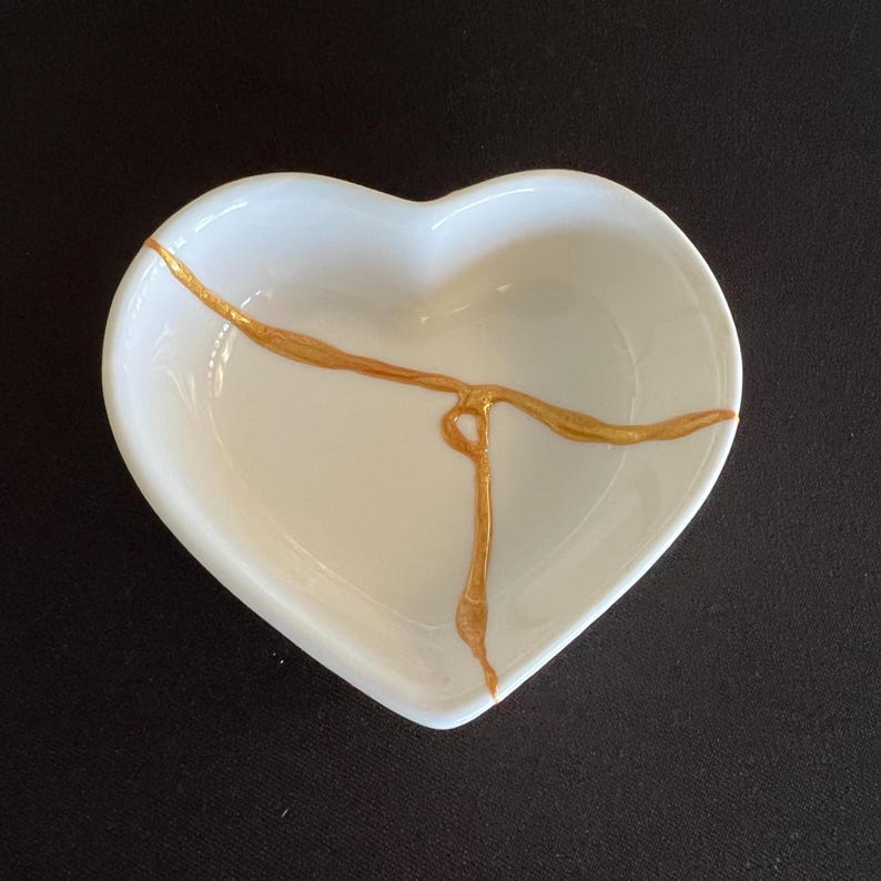 May include: A white heart-shaped ceramic dish featuring a prominent gold-coloured crack repair line that extends across the interior surface, mimicking the kintsugi technique with a smooth, glossy finish and a deep, concave centre design.