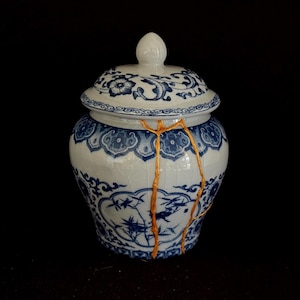 Peut inclure: Un pot en céramique blanc et bleu avec un couvercle, orné de motifs floraux et de bambous. Le pot présente des réparations kintsugi dorées. Fond noir.