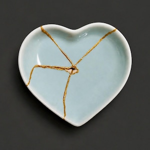 May include: A heart-shaped ceramic dish in a pale blue glaze featuring a kintsugi-style repair with prominent gold-coloured lines radiating from the centre, highlighting the cracks across the smooth, glossy surface of the small trinket bowl.
