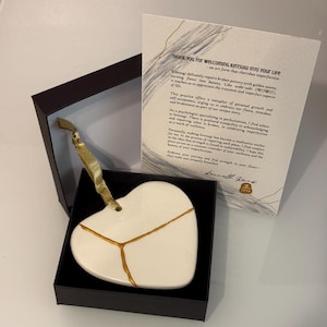 Może przedstawiać: Biała ceramiczna ozdoba w kształcie serca ze złotymi naprawami w stylu kintsugi, prezentowana w ciemnym pudełku ze złotą wstążką. Dołączona jest również karta z tekstem o kintsugi, japońskiej sztuce naprawy potłuczonej ceramiki.