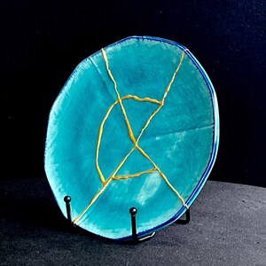 May include: A turquoise ceramic plate with gold-colored kintsugi repair lines. The plate has a dark blue rim and is displayed on a black metal stand. The background is dark.
