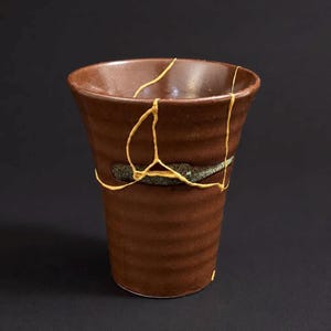May include: A brown ceramic cup featuring a series of horizontal ridges along its exterior surface. The cup displays visible cracks that have been repaired with a gold-colored lacquer, creating a distinct network of lines across the body.