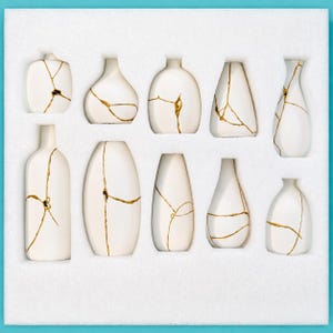 May include: A collection of nine white ceramic vases arranged in a white foam tray. Each vase features distinct gold-coloured lines resembling kintsugi repair patterns, with varying shapes including rounded, cylindrical, and tapered silhouettes.