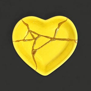 May include: A heart-shaped dish in a bright yellow finish featuring a series of intersecting cracks filled with a metallic gold-coloured material. The dish has a smooth, glossy surface and a distinct, symmetrical heart outline with a curved top.