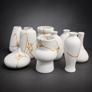 May include: A collection of nine matte white ceramic vases of varying shapes and sizes. Each vessel features distinct gold-colored lines that mimic the appearance of repaired cracks, creating a decorative pattern across the smooth white surfaces.