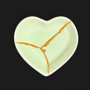 May include: A heart-shaped ceramic dish in a pale mint green colour. The dish features a prominent Y-shaped crack repair running across the centre, filled with a metallic gold-coloured material that contrasts with the smooth, light green surface.