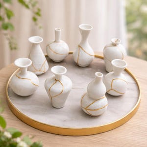 Może przedstawiać: Kolekcja ośmiu białych ceramicznych wazonów ze złotymi akcentami w stylu kintsugi. Wazony, o różnych kształtach, są ułożone na marmurowej tacy ze złotą obwódką. Zestaw stoi na drewnianym stole.