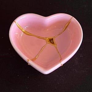 May include: A heart-shaped ceramic dish in a soft pastel pink colour. The surface of the dish features a prominent crack that has been repaired with a metallic gold finish, creating a distinct branching pattern across the centre of the bowl.