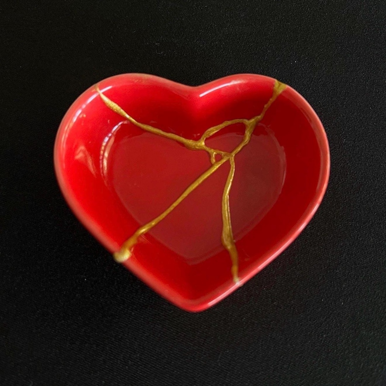 May include: A heart-shaped red ceramic dish featuring a gold-coloured crack repair pattern across its surface. The bowl has a smooth, glossy finish and a deep interior basin, with the metallic gold lines creating a distinct branching design.