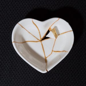 May include: A white heart-shaped ceramic dish featuring a series of cracks repaired with a metallic gold finish. The dish has a smooth, glossy surface with the gold lines radiating from the centre, highlighting the broken and reassembled pieces.