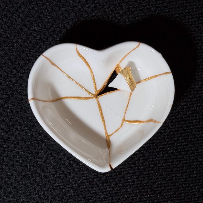 May include: A white heart-shaped ceramic dish featuring a series of cracks repaired with a metallic gold finish. The dish has a smooth, glossy surface with the gold lines radiating from the centre, highlighting the broken and reassembled pieces.