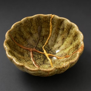 May include: A ceramic bowl with a scalloped rim and a mottled green and brown glaze. The bowl features gold-colored Kintsugi repairs, highlighting the cracks. The bowl is a decorative piece.