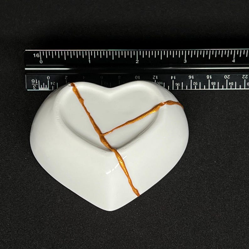 May include: A white heart-shaped ceramic dish featuring gold-coloured repair lines across the surface. A black ruler is positioned above the dish, showing a width of approximately 9 centimetres for the heart-shaped object.