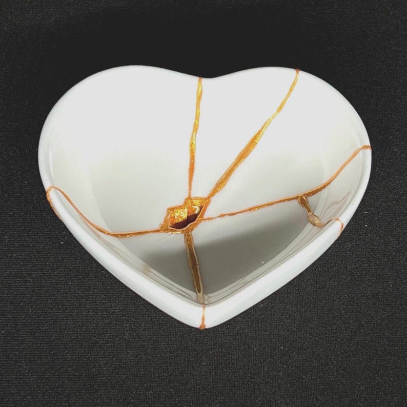 May include: A white heart-shaped ceramic bowl featuring a kintsugi-style repair with gold-coloured lines radiating from the centre. The smooth, glossy surface of the bowl is contrasted by the textured, metallic gold veins that connect the cracks.