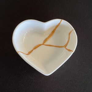 May include: A white heart-shaped ceramic dish featuring a prominent gold-coloured crack repair design across the centre. The bowl has smooth, curved edges and a glossy finish, with the metallic gold lines mimicking the kintsugi repair technique.