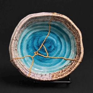 May include: A ceramic bowl with a unique kintsugi repair. The bowl features a vibrant turquoise center with a spiral design, surrounded by a textured rim. Gold-colored epoxy fills the cracks, creating a striking contrast. The bowl is displayed on a black stand.